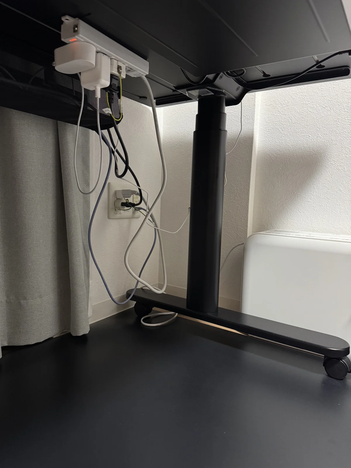 COFO Desk Studioの天板裏まわり。脚ベースのキャスターと、壁のコンセントに直挿しした電源プラグ、周辺機器用の電源タップが写っている